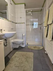 Ein modernes Badezimmer mit weißen Fliesen und einer großen Dusche. Es gibt ein Waschbecken, eine Toilette und weiche Badezimmermatten in neutralen Farben.