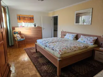 Ein gemütliches Schlafzimmer mit einem großen Bett und bunten Bettwäsche. Der Raum ist hell und freundlich, mit einer Sitzgelegenheit und einem schönen Teppich.