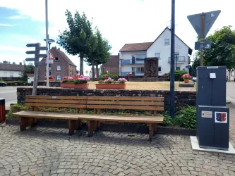 Eine ruhige Straßenszene mit einer Holzbank, einem Trinkwasserbrunnen und bunten Blumenbeeten. Im Hintergrund sind typische Häuser und Wegweiser zu sehen.