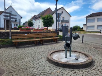 Ein eleganter Brunnen steht auf einem kleinen Platz mit gepflastertem Boden. Im Hintergrund sind schmucke Häuser und blühende Blumen in Kübeln zu sehen.