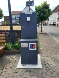 Ein grauer Trinkwasserbrunnen steht an einer Straße in einem Wohngebiet. Im Hintergrund sind Häuser und Bäume zu sehen.