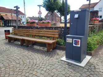Eine Holzbank steht auf einem gepflasterten Platz. Neben der Bank befindet sich ein grauer Trinkwasserbrunnen.
