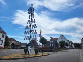 Ein schöner, geschmückter Maibaum steht im Zentrum einer kleinen Gemeinde. Der Himmel ist blau und es gibt einige Wolken.