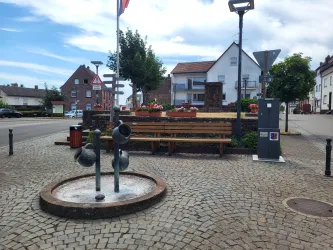 Ein ruhiger Platz mit einer Bank, einem Brunnen und Blumenbeeten. Im Hintergrund sind Häuser und Verkehrsschilder sichtbar.