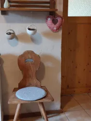 Ein kleiner Holzstuhl mit einer runden Sitzfläche und einem herzförmigen Dekorationsobjekt an der Wand. Im Hintergrund hängen kleine Töpfe mit Pflanzen.