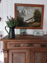Ein Holzschrank mit einer dekorativen Blume in einer schwarzen Kanne und einem Bilderrahmen mit einer Landschaftsmalerei. Im Hintergrund sind ein Glasobjekt und eine kleine Box zu sehen.