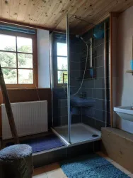 Ein helles Badezimmer mit einer gläsernen Dusche und großen Fenstern. Die Wände sind mit Fliesen verkleidet und es gibt einen Waschtisch sowie einen Heizkörper.