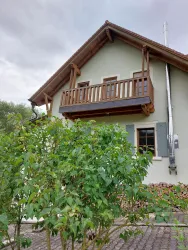 Ein charmantes Haus mit einem Balkon und Fensterläden. Im Vordergrund sind grüne Sträucher zu sehen.