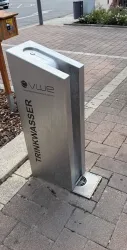 Ein moderner Trinkwasserbrunnen aus Edelstahl steht auf einem gepflasterten Gehweg. Daneben sind einige Blumen und eine Straße sichtbar.