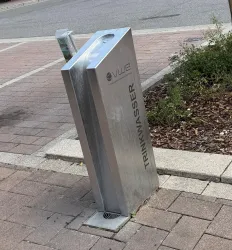 Ein moderner Wasserspender aus Edelstahl steht auf einem Bürgersteig. Darauf steht das Wort "TRINKWASSER".