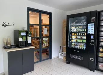 Ein heller Raum mit einem Snackautomaten und einer Kaffeemaschine. Im Hintergrund sieht man eine Tür, die zu einem anderen Bereich führt.