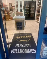 Ein einladender Eingang zu einem Laden mit einem willkommen heißenden Teppich. Im Hintergrund sind Regale mit Lebensmitteln zu sehen.