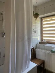 Ein Badezimmer mit einer Duschvorhang vor einer Badewanne. Hinter dem Vorhang sind Fliesen und ein Fenster mit Rollläden zu sehen.