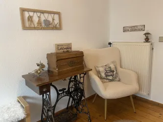 Ein gemütlicher Wohnbereich mit einem beigen Sessel und einem alten Nähmaschinen Tisch. An der Wand hängen dekorative Elemente und ein Schild mit einer kurzen Aufschrift.