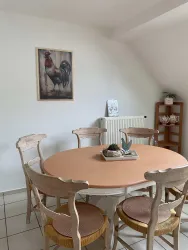 Ein gemütlicher Essbereich mit einem runden Tisch und Holzstühlen. An der Wand hängt ein Bild eines Huhns, und im Hintergrund steht ein Regal mit Tassen.