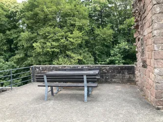 Ein Platz mit einem Holztisch und Bänken steht vor einer grünen Baumlandschaft. Die Umgebung wirkt ruhig und einladend.