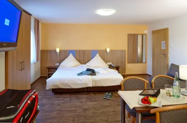 Ein gemütliches Hotelzimmer mit einem großen Doppelbett und modernem Mobiliar. Neben dem Bett befindet sich ein Tisch mit Snacks und Getränken.
Das Bild zeigt unser barrierefreies Appartement.