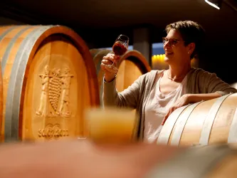Eine Frau genießt ein Glas Wein in einem Weinkeller. Im Hintergrund sind große Holzfässer sichtbar.