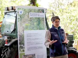 Eine Frau spricht neben einem Traktor und einem Informationsplakat. Das Plakat thematisiert die Begrünung im Ökosystem Weinberg.
