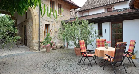 Ein einladender Innenhof mit gemütlichen Stühlen und einem Tisch. Umgeben von Pflanzen und historischen Gebäuden.