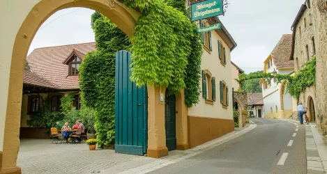 Eine malerische Straße mit Gebäuden und vielen Pflanzen. Im Hintergrund sitzen Menschen an einem Tisch und genießen die Atmosphäre.