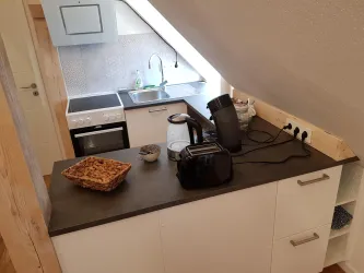 Eine moderne Küchenecke unter dem Dach mit einer Kochplatte, einem Wasserkocher und einem Toaster. Der Raum wirkt hell und freundlich mit Holzdetails und einer praktischen Arbeitsfläche.