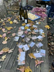 Ein rustikaler Holztisch ist mit bunten Herbstblättern, Weingläsern und einer Platte mit Keksen dekoriert. Im Hintergrund sind einige Flaschen Wein zu sehen.