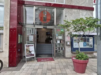 Ein modernes Gebäude mit einer gläsernen Eingangstür. Vor der Tür steht ein roter Blumentopf mit einer Pflanze.
