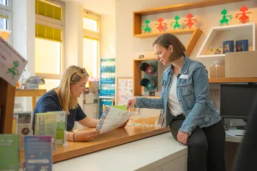Zwei Frauen stehen in einem Empfangsbereich. Eine Frau liest ein Dokument, während die andere sie aufmerksam beobachtet.