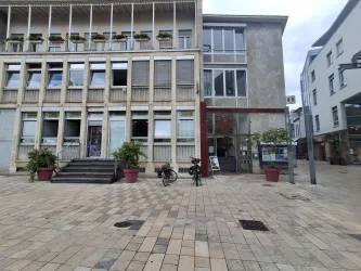 Ein Gebäude mit einem modernen Erscheinungsbild und großen Fenstern. Vor dem Gebäude befindet sich ein Platz mit Pflanzkübeln und Fahrrädern.