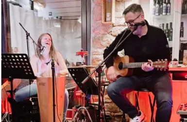 Eine Sängerin und ein Gitarrist spielen live in einem gemütlichen Ambiente. Die Atmosphäre ist entspannt und kreativ, mit Musik-Equipment im Hintergrund.