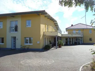 Ein modernes, gelb gestrichenes Gebäude mit mehreren Eingängen. Der gepflasterte Hof ist sauber und einladend, umgeben von Pflanzen.