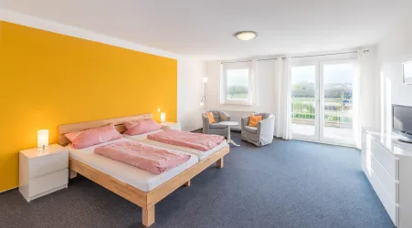 Ein modernes Schlafzimmer mit einem großen Bett und einer leuchtend gelben Wand. Große Fenster sorgen für viel Tageslicht und einen Blick ins Freie.