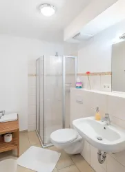 Ein modernes Badezimmer mit einer Duschkabine und einem Waschbecken. Auf der linken Seite steht ein Holzregal mit Toilettenartikeln.