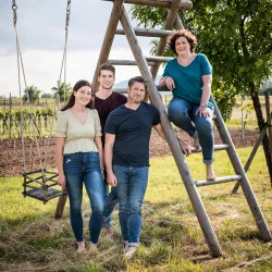 Eine Gruppe von vier Personen steht vor einer Schaukel und einer Holzleiter im Freien. Die Umgebung zeigt grüne Felder und Bäume.