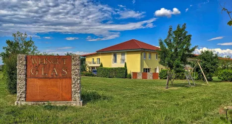 Ein schönes gelbes Haus mit einem roten Dach und grünem Garten. Im Vordergrund steht ein Schild mit der Aufschrift "WEINGUT GLAS".