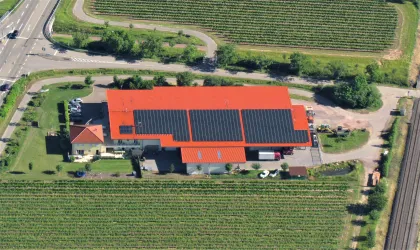 Ein modernes Gebäude mit einem roten Dach und Solarpanelen ist umgeben von Weinbergen. In der Nähe verläuft eine Straße.