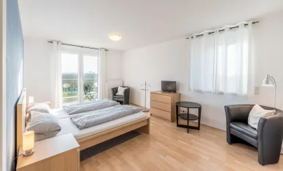 Ein helles Schlafzimmer mit einem komfortablen Bett und modernen Möbeln. Große Fenster sorgen für viel Tageslicht und bieten einen Blick ins Grüne.