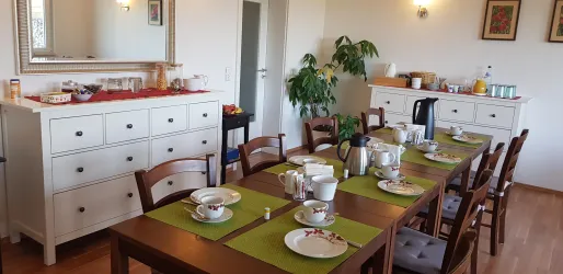 Ein einladender Speisesaal mit einem langen Holztisch und gedeckten Tellern. Im Hintergrund stehen eine Kommode und verschiedene Leckereien bereit.