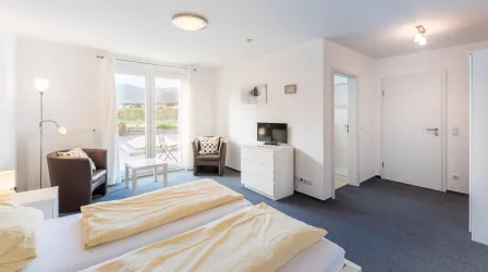 Ein helles Schlafzimmer mit einem großen Fenster und Blick nach draußen. Es gibt ein Bett, einen Fernseher und gemütliche Sitzgelegenheiten.