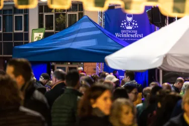 Ein lebendiges Weinfest mit vielen Menschen und einem blauen Zelt. Im Hintergrund ist ein Banner mit dem Schriftzug „Deutsches Weinfest“ sichtbar.