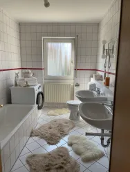 Ein modernes Badezimmer mit einer Badewanne, einem Waschbecken und einer Toilette. Weiche Teppiche auf dem Boden und viel Tageslicht durch das Fenster.