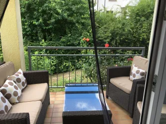 Ein gemütlicher Balkon mit einem Tisch und zwei Sitzen aus Rattan. Im Hintergrund sieht man üppiges Grün und ein paar blühende Blumen.