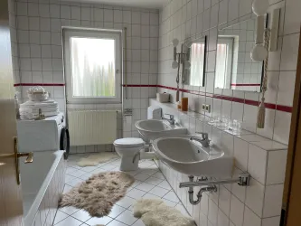 Ein modernes Badezimmer mit weißen Fliesen und einem großen Fenster. Es gibt ein Waschbecken, eine Toilette und einen Wäscheautomaten, sowie weiche Teppiche auf dem Boden.