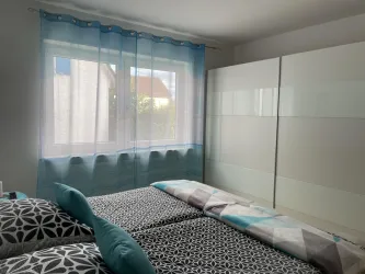Ein gemütliches Schlafzimmer mit zwei Betten und bunten Kissen. Große Fenster mit Vorhängen lassen viel Licht herein.
