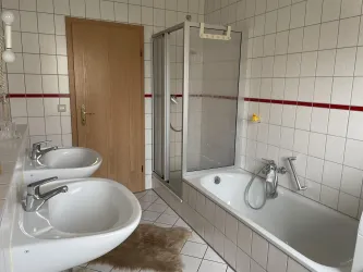 Ein modernes Badezimmer mit zwei Waschbecken, einer Dusche und einer Badewanne. Die Wände sind mit hellen Kacheln und einem roten Streifen dekoriert.