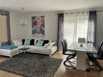 Ein modernes Wohnzimmer mit einem weißen Sofa und einem eleganten Esstisch. Große Fenster sorgen für viel Tageslicht und eine einladende Atmosphäre.