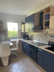 Eine moderne Küche mit blauen Schränken und hellen Wänden. Am Tisch steht ein weißer Stuhl und ein Fenster bietet Ausblick ins Freie.
