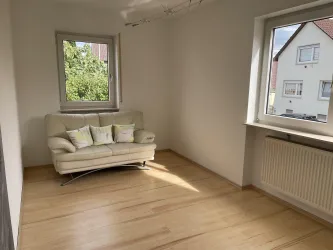 Ein heller Raum mit einem bequemen weißen Sofa und drei Kissen. Das Fenster bietet einen Blick auf die grünen Bäume draußen.