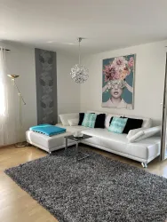 Ein modernes Wohnzimmer mit einem weißen Sofa und bunten Kissen. An der Wand hängt ein KUNSTWERK mit einer Schönheit, die Blumen trägt.
